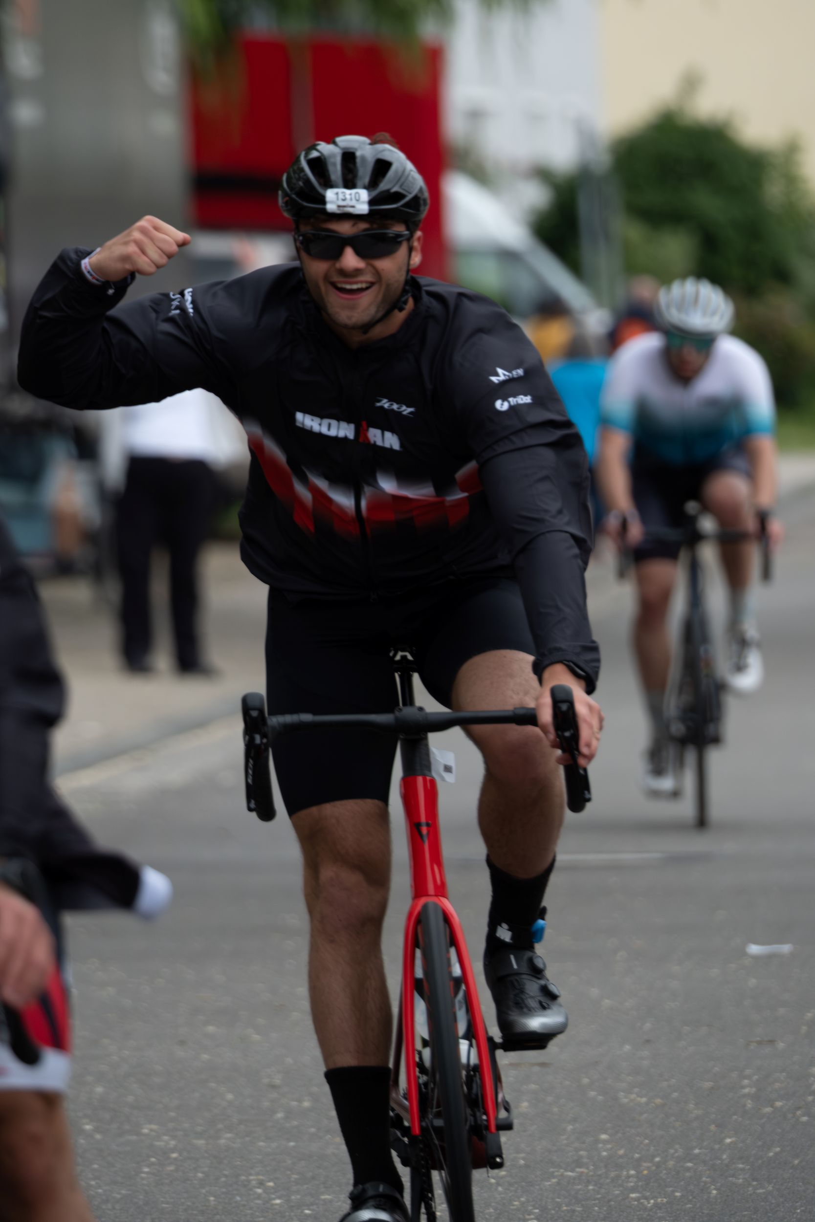 Twan op rode racefiets tijdens halve Ironman, flexed biceps en kijkt lachend naar camera — het resultaat van consistente hybride training.